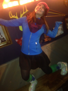 A woman in a blue jacket, pink hat, and black skirt poses with arms outstretched and one leg kicked out.