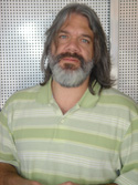 A man with long brown hair and a grey beard wearing a green striped polo shirt.