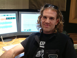 A man with wavy hair smiling in front of two computer monitors in a dark room.