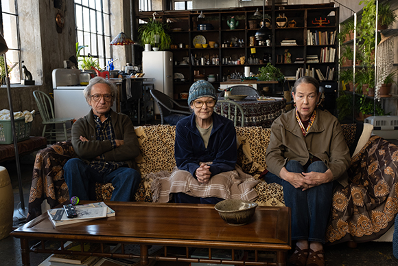 Three people sit on a patterned sofa in a room filled with books, plants, and various household items.