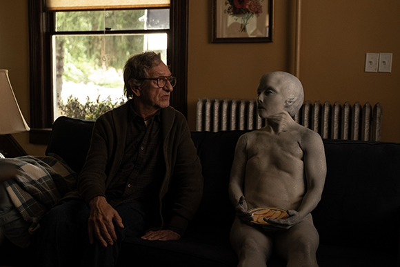 An older man with glasses looks at a grey-skinned alien sitting on a dark sofa in a dimly lit room.