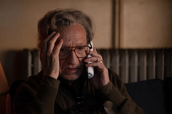 An older man with wavy grey hair wearing glasses, holding his head in one hand and a phone to his ear with the other.