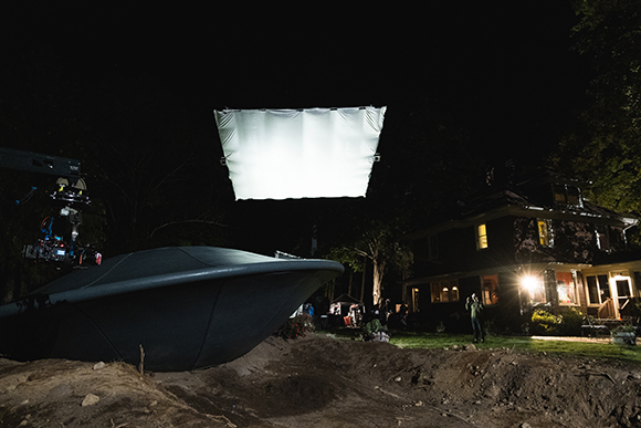 A large white light panel hangs in the night sky above a dark, saucer-shaped object and a house.