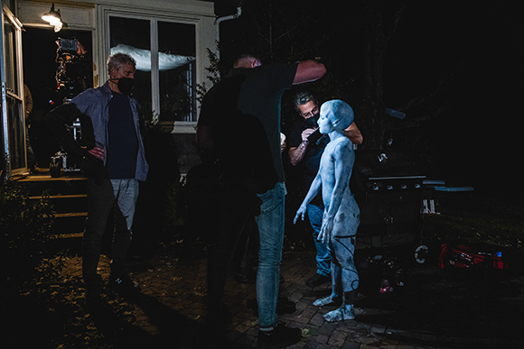 Film crew members surround a blue-skinned alien creature on a dark outdoor set at night.