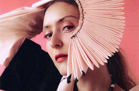 A woman with pearl eye makeup wearing a large, fan-like pink pleated headpiece against a pink background.