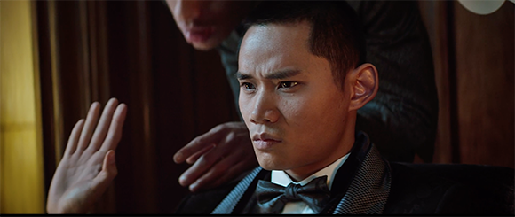 A man in a black tuxedo and bowtie looks forward with a serious expression.