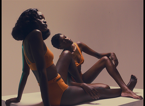 Two Black women in orange swimwear posing artistically on a white platform against a tan background.