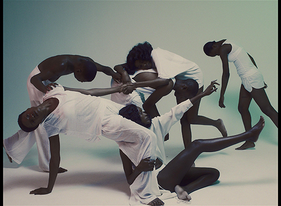 Six people in white clothing perform expressive, sculptural dance movements against a plain background.