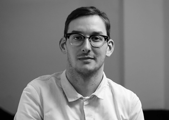 A man with short dark hair and glasses wearing a light-colored collared shirt.