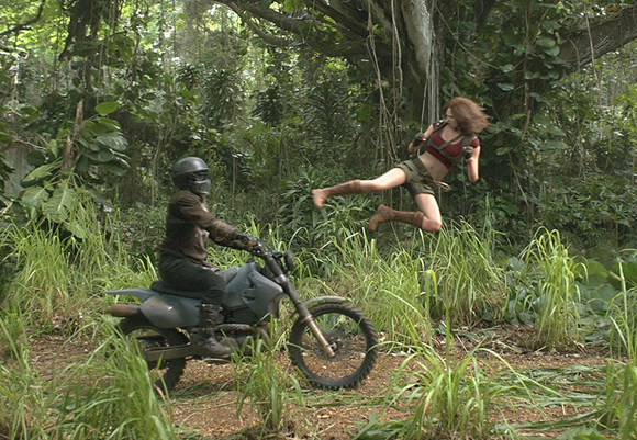 A woman in a red top jumps mid-air in a lush forest near a man riding a grey motorcycle.