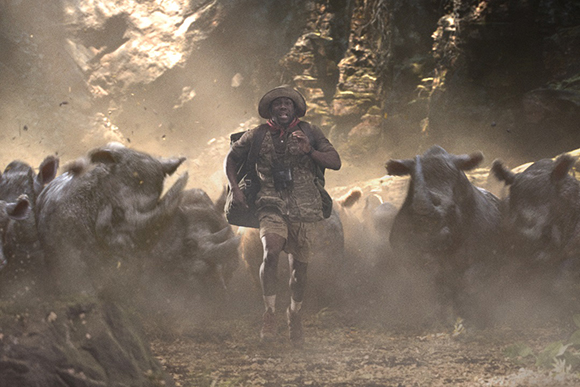 A man in safari gear runs toward the camera through dust, pursued by several large warthogs.