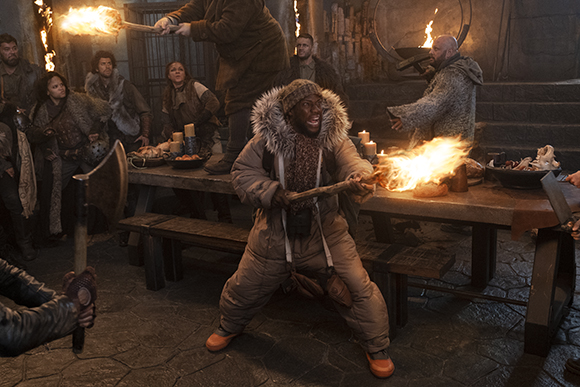 A man in a tan parka holds a flaming torch while surrounded by people in a dimly lit stone hall.