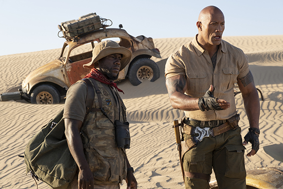 A man in a tan shirt gestures while another man in a sun hat stands beside a rusted desert vehicle.
