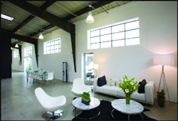 A bright, modern loft with white walls, high ceilings, large windows, a white sofa, and marble coffee tables.