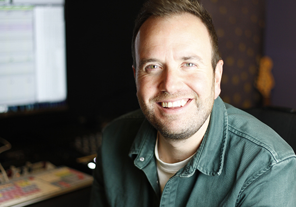 A smiling man with short brown hair and light stubble wearing a green button-down shirt.