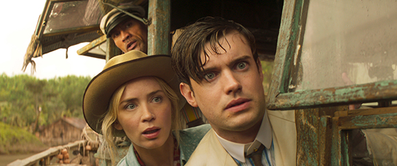 A blonde woman in a wide-brimmed hat and a man in a light suit look on with wide eyes and surprised expressions.