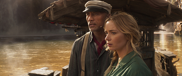 A man in a cap and a woman with blonde hair look toward the distance from a wooden boat on water.