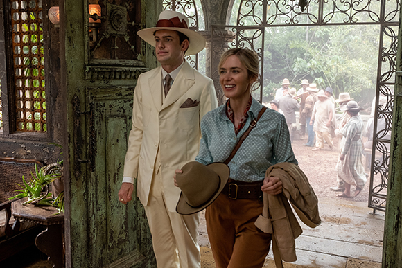 A man in a cream suit and hat walks beside a woman in a blue shirt and brown trousers through a metal gate.