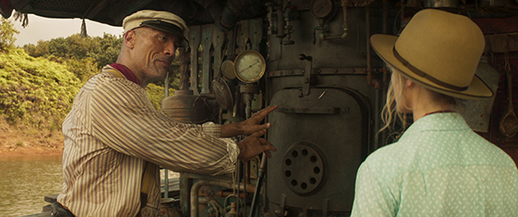 A man in a cap gestures toward complex machinery while speaking to a woman in a wide-brimmed hat.