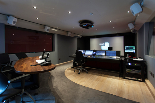 A dark control room featuring a large mixing console, multiple computer monitors, and a projector screen.