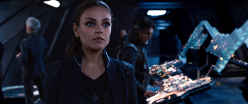 A woman with dark hair looks thoughtfully into the distance in a dark, high-tech environment.