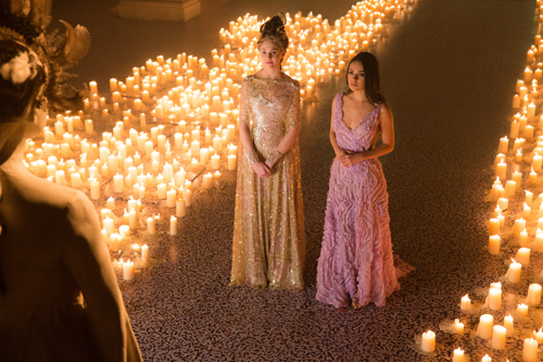 Two women in long evening gowns stand in a room where hundreds of lit candles cover the floor.