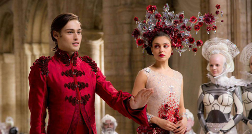 A man in a red military-style jacket and a woman in a floral headpiece stand in a stone hall.