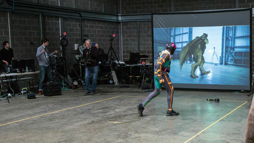 A person in a motion capture suit performs in front of a large screen showing a digital creature.