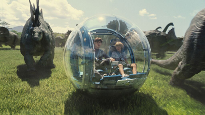Two children sit inside a clear spherical vehicle in a grassy field surrounded by large dinosaurs.