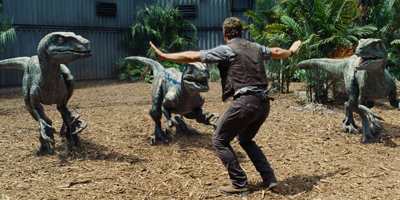 A man in a vest stands with arms wide, facing three velociraptors in a wooded enclosure.