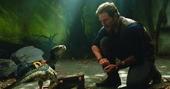 A man in a brown vest kneels on the ground, looking intently at a small blue and grey dinosaur.