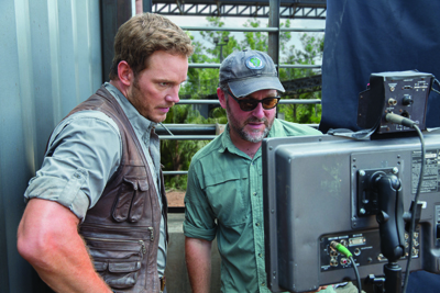 A man in a brown vest and a man in a grey cap look at a large video monitor outdoors.