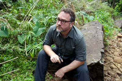 A man with glasses and a beard sits on a large rock amidst dense green foliage.