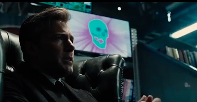 A man in a dark suit sits in a leather chair looking toward a screen with a teal skull icon.