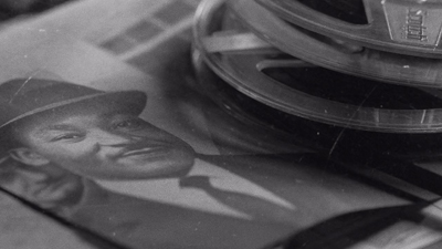 Black and white close-up of a man's portrait in a hat next to stacked film reels.