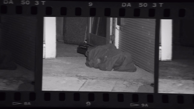 Black and white film frame of a person lying face down on a sidewalk next to a closed metal shutter.