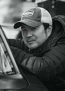 Black and white portrait of a man wearing a baseball cap and a quilted jacket leaning on a vehicle.