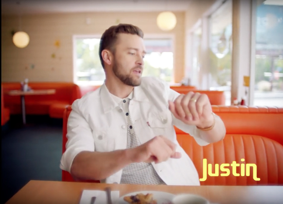 A man with a beard wearing a white denim jacket sits at a table in a diner with orange booths.