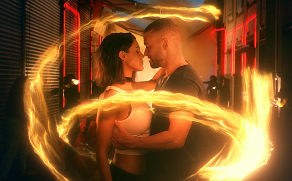 A man and woman embrace in a dark alleyway surrounded by swirling, glowing golden light trails.