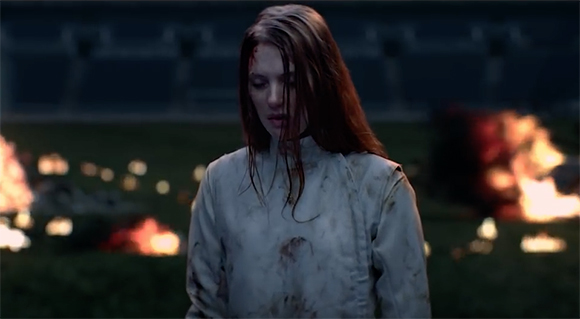 A woman with long reddish hair and blood on her face and clothing stands in a dark field with fires behind her.