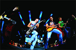 Rock band members playing electric guitars and bass on a dark stage with blue light beams.