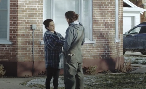 Two people stand facing each other on a lawn in front of a brick house.