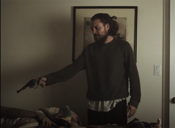 A man with long hair and a beard points a handgun toward a person lying in a bed.
