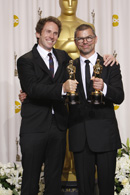 Two men in black suits smiling and holding gold Oscar awards against a white backdrop.