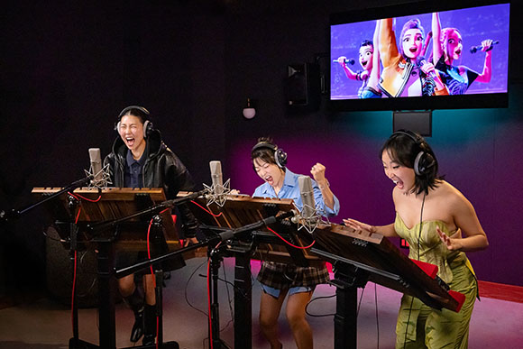 Three young women singing and playing electronic keyboards in a dark music studio.