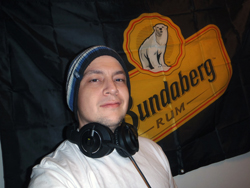 Man wearing headphones and a beanie in front of a black flag with a yellow Sundberg Rum logo featuring a polar bear.