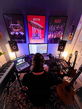 A person sits at a desk with a computer, MIDI keyboard, and guitars, under rock band posters.