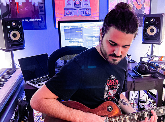 A man with dark hair in a bun plays an electric guitar in a studio with monitors and a computer.