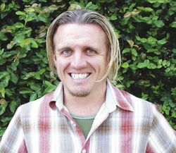 A smiling man with shoulder-length blonde hair wearing a plaid shirt in front of green foliage.