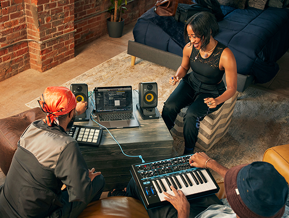 People using a laptop, speakers, and a MIDI keyboard in a room with brick walls.